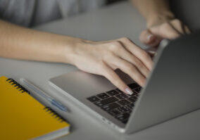 3-admin0Writers Close-up of a person's hands typing on a silver laptop keyboard next to a yellow spiral notebook and a blue pen.