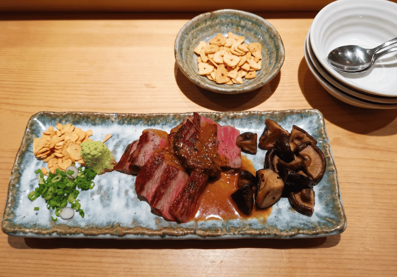 A rectangular blue and white plate featuring sliced medium-rare wagyu steak topped with a savory sauce, served with sauteed shiitake mushrooms, chopped scallions, wasabi, and a side of crispy garlic chips.