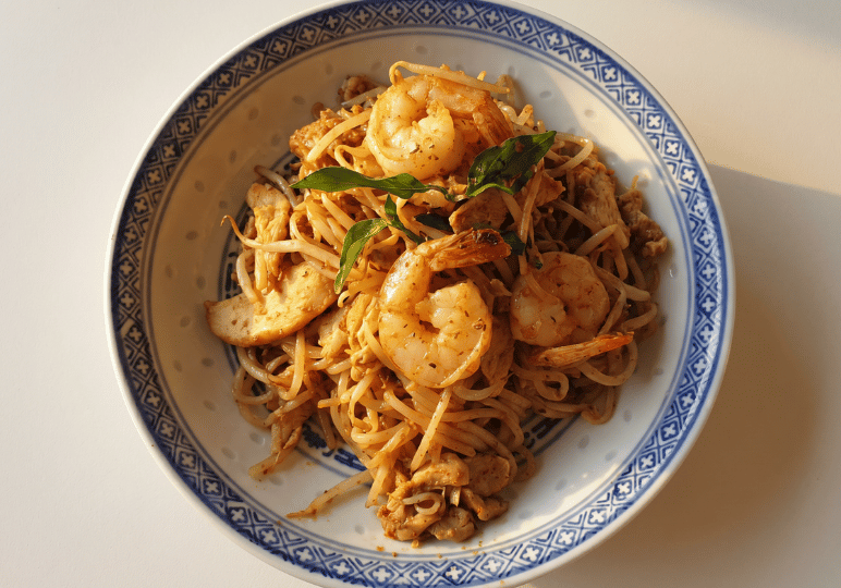 3-curry-times-velocity A top-down view of a bowl of stir-fried noodles topped with large prawns, chicken pieces, and bean sprouts.