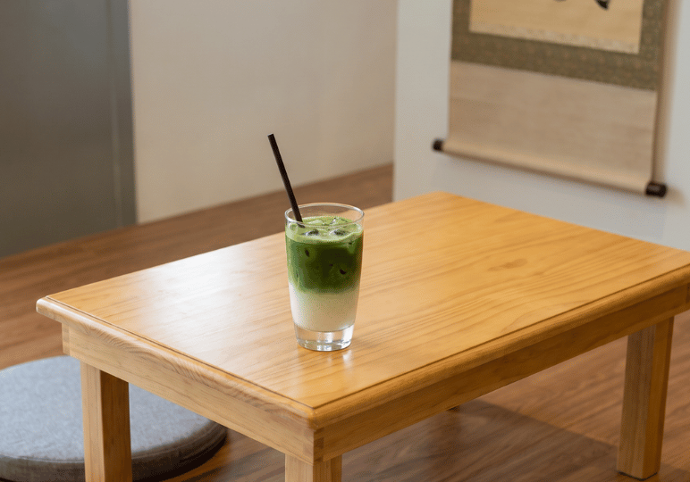 A glass of iced matcha latte showing distinct layers of green matcha and white milk, sitting on a low wooden table beside a grey floor cushion.