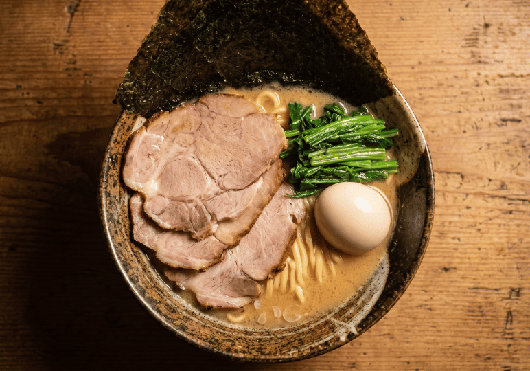 3-orchard-plaza-stalls Top-down view of a rich bowl of Japanese ramen topped with thick slices of chashu pork, a soft-boiled egg, fresh spinach, and a sheet of nori on a rustic wooden table.