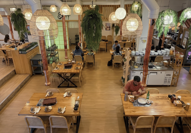 3-restaurant-interior An airy restaurant interior with light wood dining tables, woven rattan lampshades, hanging copper exhaust pipes, and lush green ferns draped from the ceiling to create a fresh, natural aesthetic.