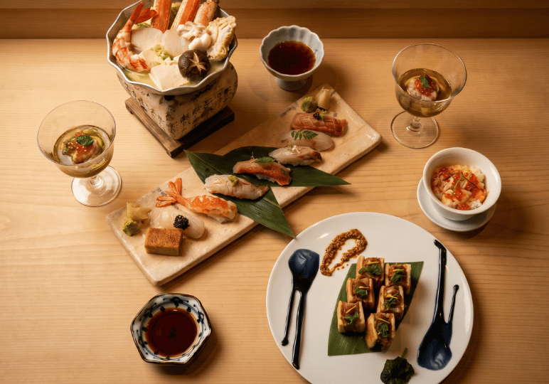 A high-angle spread of an upscale Japanese meal, featuring a hot pot with crab legs and mushrooms, assorted platters of premium nigiri sushi, and small traditional side dishes on a light wood table.