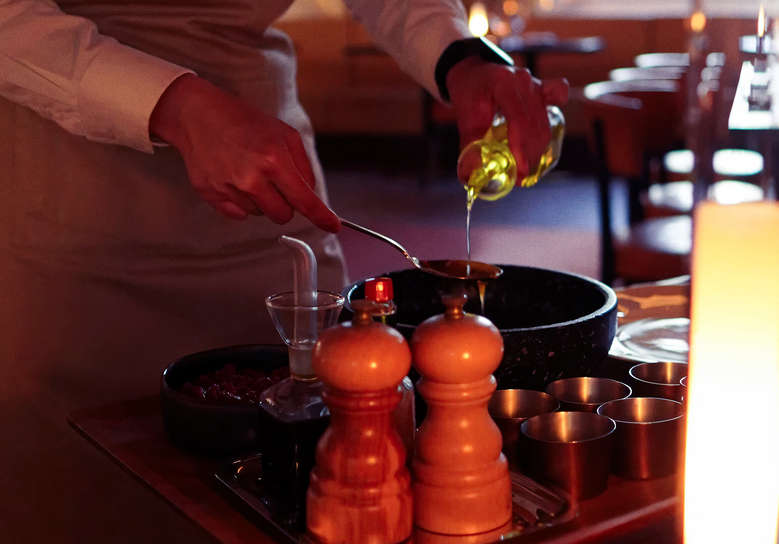 Tableside culinary service showing a server in an apron drizzling oil into a spoon above a large black bowl with ingredients and utensils on the table.