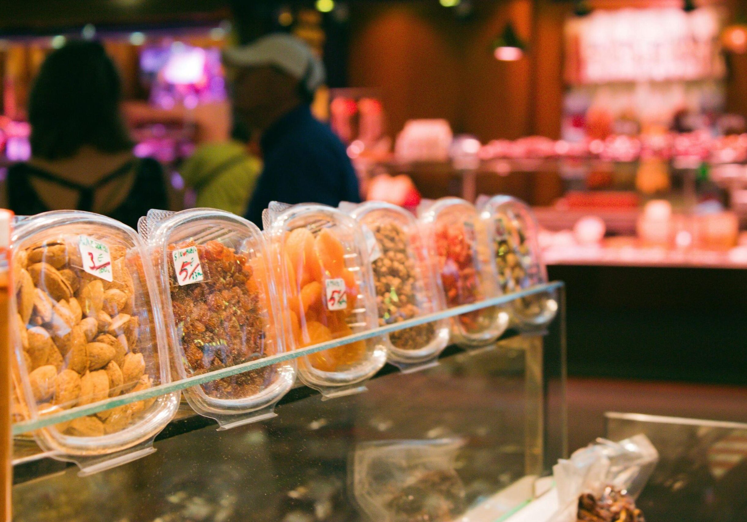 This image displays a row of pre-packaged snacks, including almonds, raisins, and dried apricots, neatly arranged on a glass display shelf in a market setting. The background is softly blurred, showing the warm lighting and busy atmosphere of a grocery store or food stall with customers nearby.