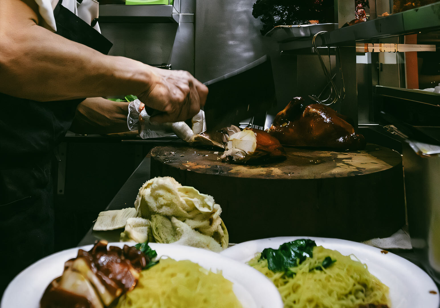 Carving roasted meat on a large wooden chopping block in a kitchen, with plates of noodles and greens prepared in the foreground.