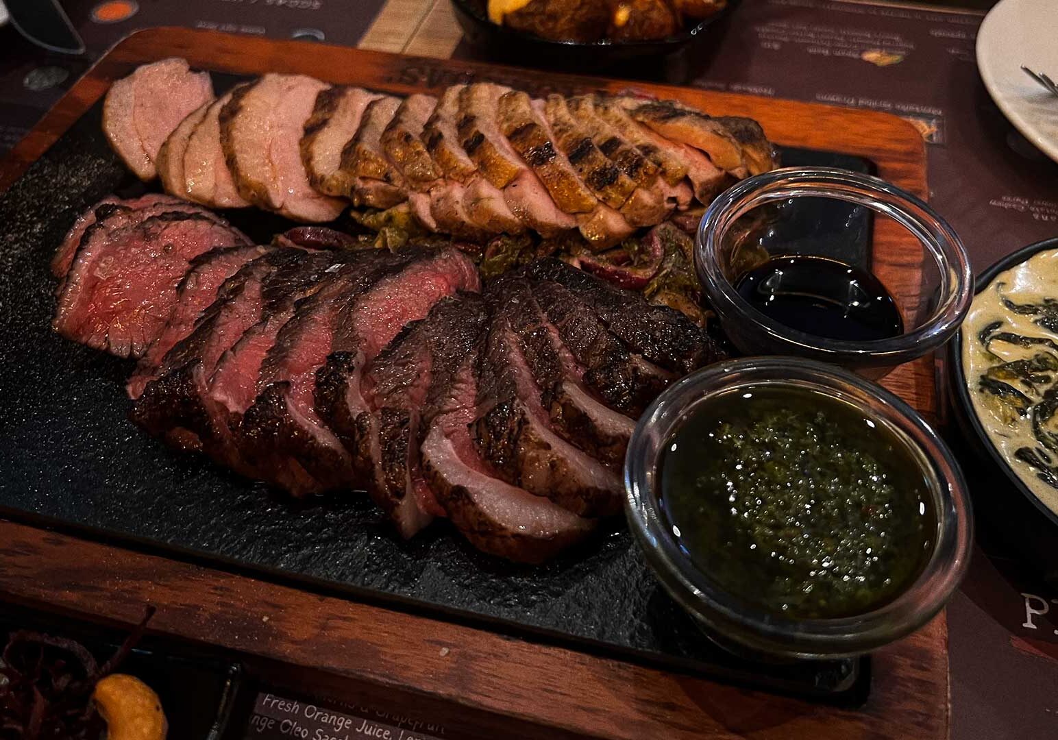 Wooden serving board with sliced grilled steak and duck breast, paired with soy-based sauce, chimichurri, and creamy spinach side dish in restaurant setting.