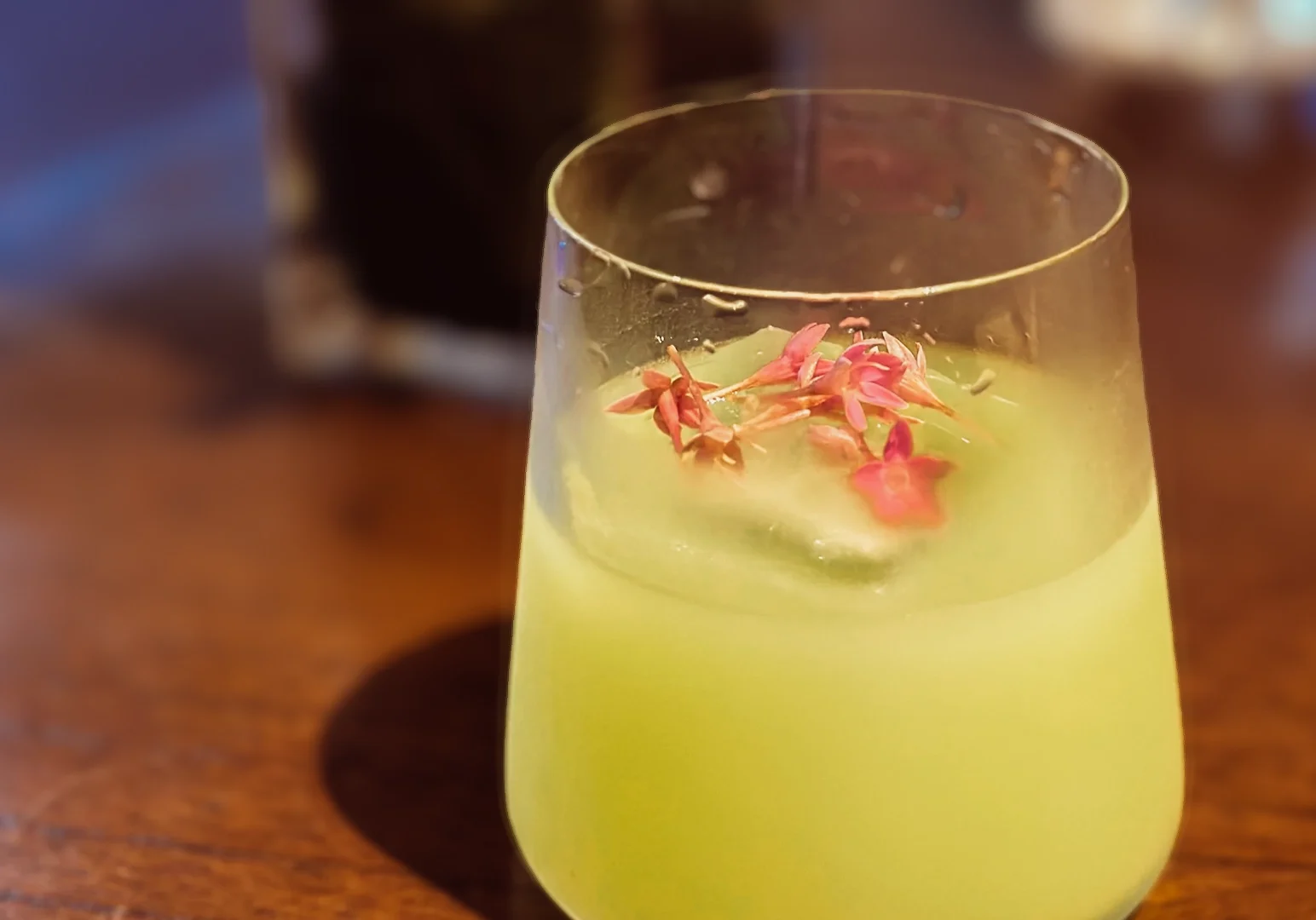4 Jiak Kim House Singapore Glass of green craft cocktail garnished with pink edible flowers and served over a large ice cube on a wooden table.