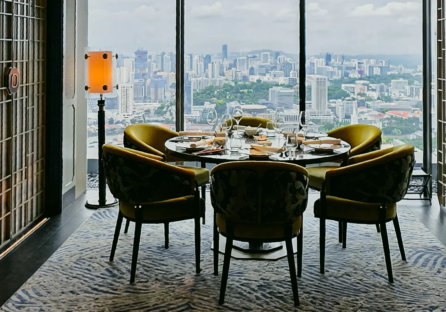 4 Jin Ting Wan Marina Bay Sands Eye‑level wide shot of a circular dining table at Jin Ting Wan Marina Bay Sands, set with fine tableware and plush chairs, framed by floor‑to‑ceiling windows overlooking the Singapore city skyline from Level 55.