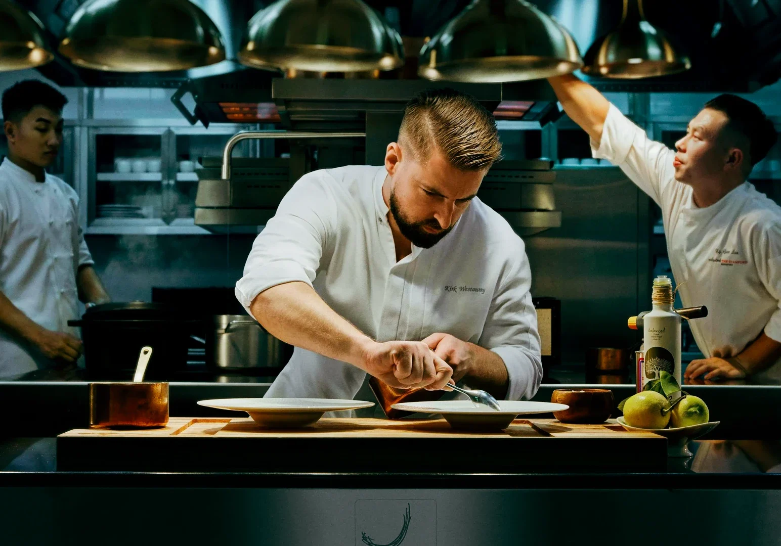 4 Kirk Westaway Chef Kirk Westaway in a modern professional kitchen carefully plating a dish under warm overhead lights, with cookware, ingredients, and kitchen staff working in the background.