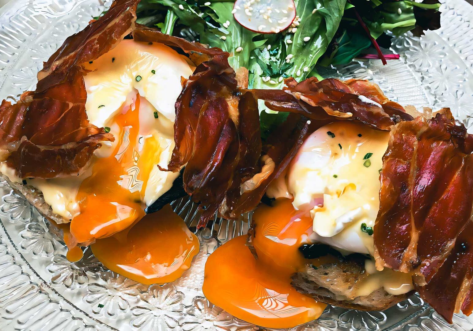 Close-up of gourmet eggs Benedict topped with crispy prosciutto, runny egg yolks, and hollandaise sauce served on toasted bread with a fresh salad on a decorative glass plate.