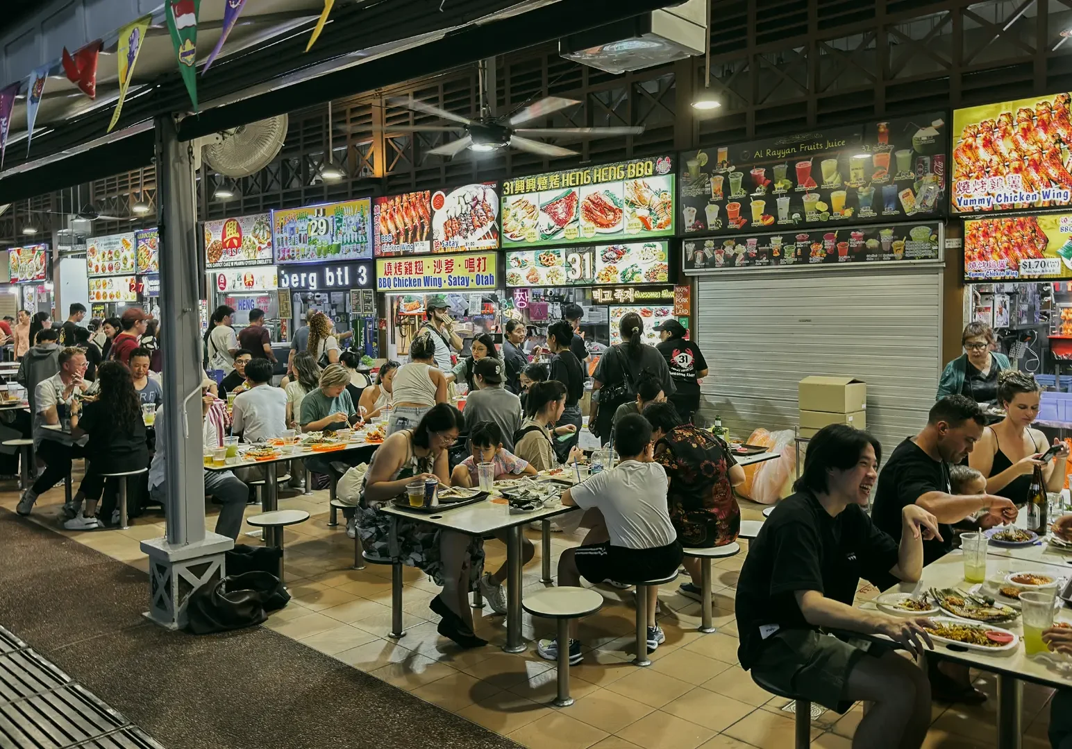 Wide-angle street-level shot of a busy night hawker centre with diners seated at long tables, illuminated food stalls, and lively local atmosphere.