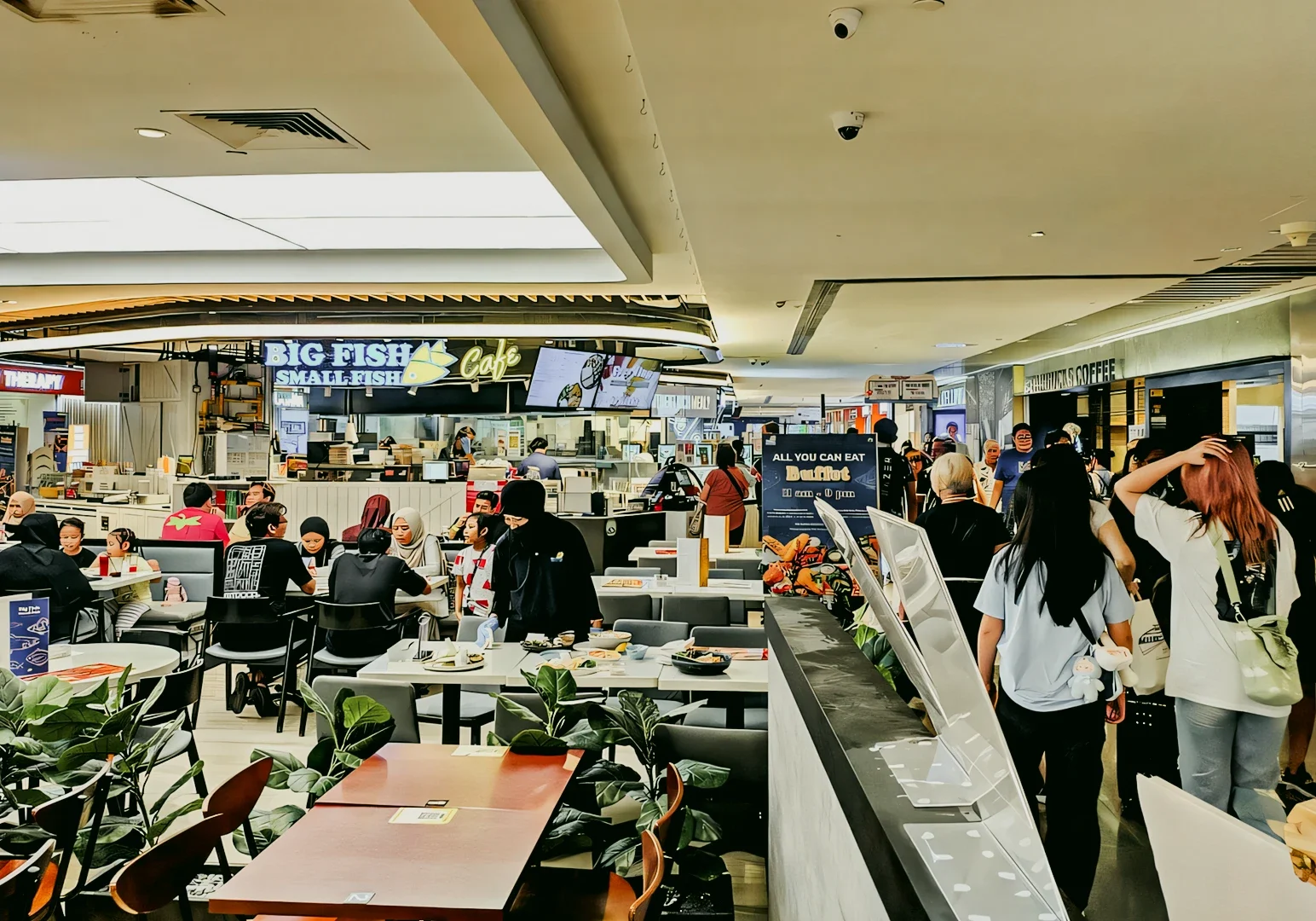 Wide-angle eye-level shot of a busy shopping mall food court with multiple dining tables, seated patrons, illuminated restaurant signage, and walkways filled with people.