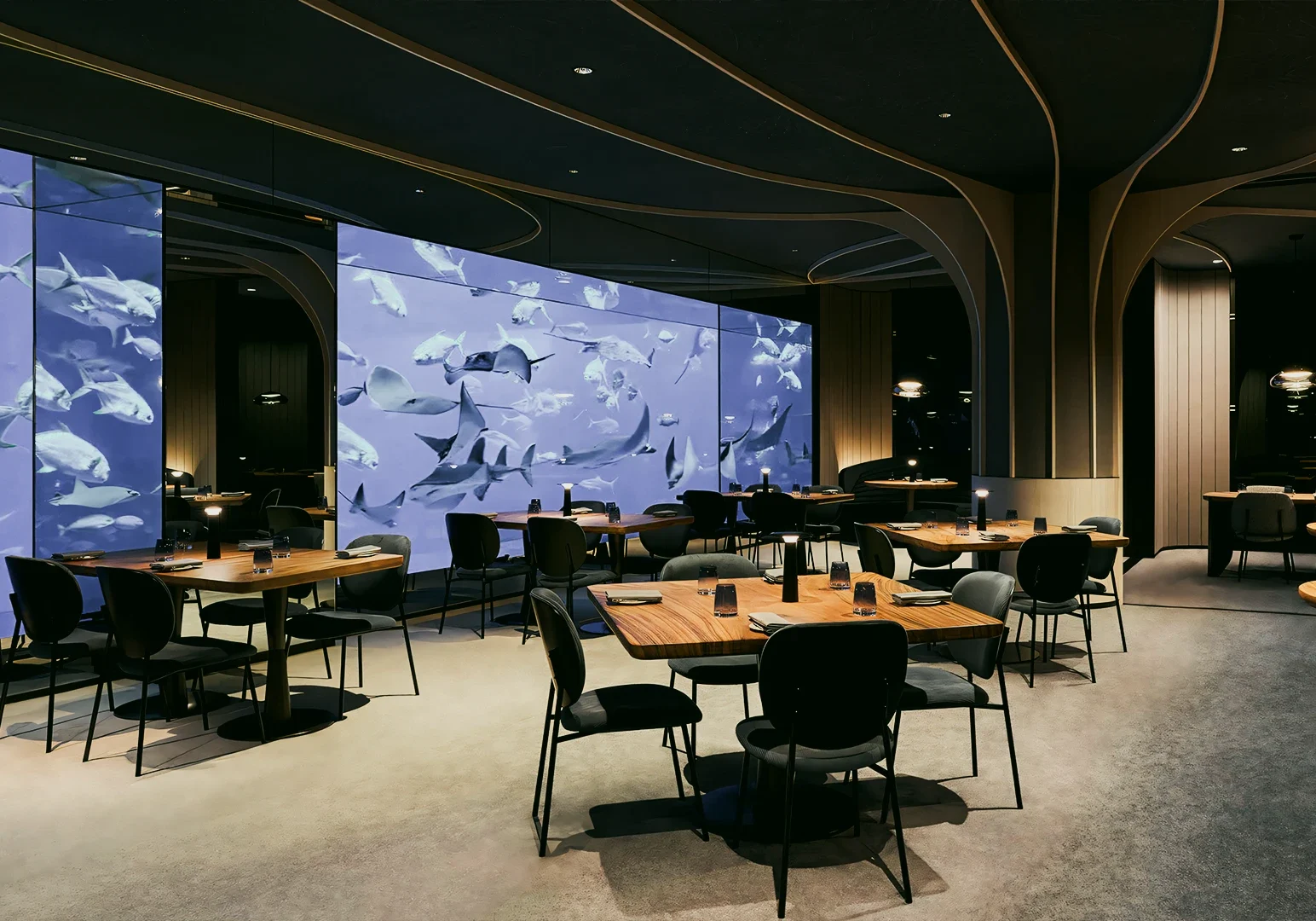 Wide‑angle interior shot of a modern restaurant dining room featuring wooden tables, ambient lighting, and a large digital aquarium wall display.