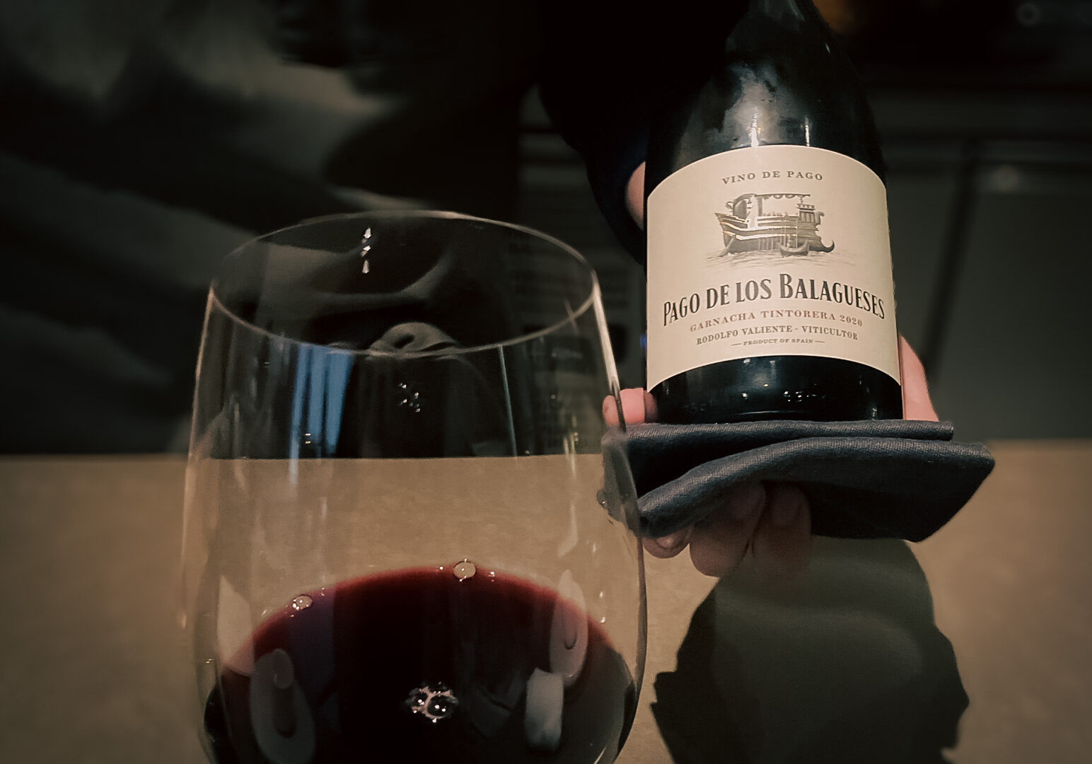 Close‑up of a glass of red wine beside a hand presenting a bottle labeled ‘Pago de los Balagueses’ in a dimly lit restaurant setting.
