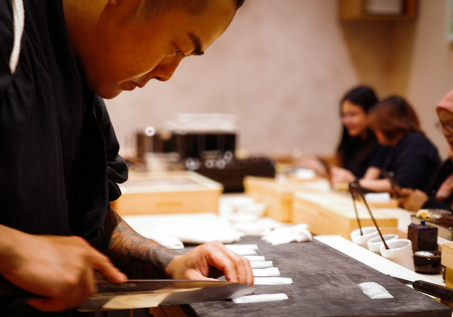 Japanese sushi chef slicing fresh white fish sashimi on dark cutting board, customers seated at counter in background.