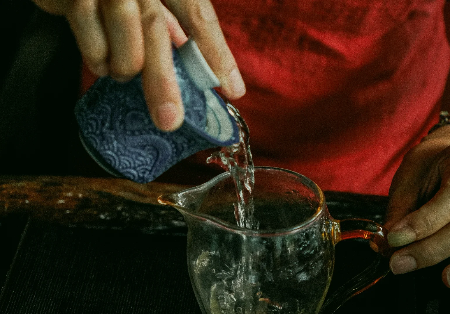 Pouring hot water into glass pitcher during Chinese tea ceremony at Tea Room by Ki-setsu – close-up action shot with slight overhead angle capturing hands-on tea preparation and mindfulness experience