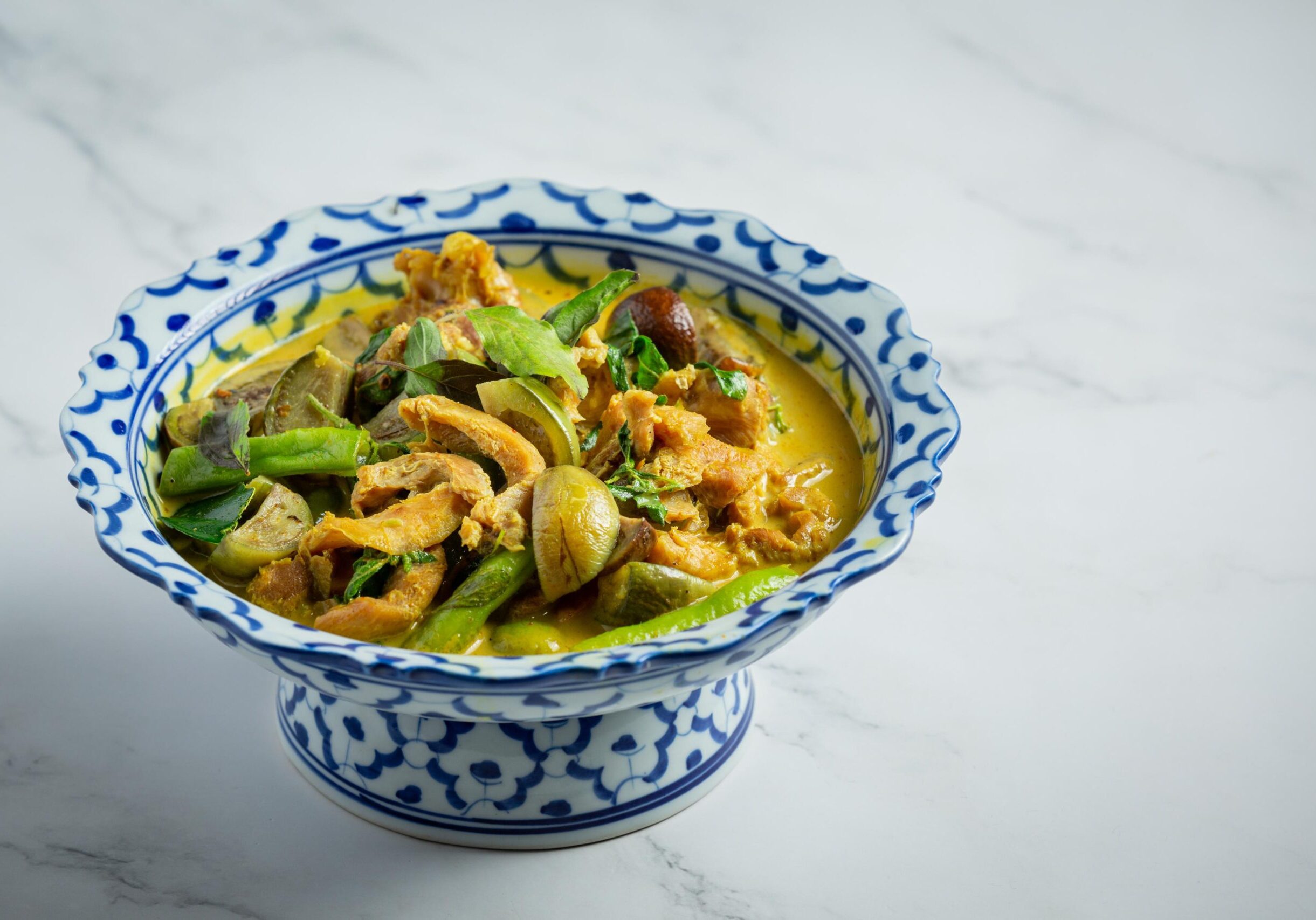 This image features a bowl of Thai green curry filled with chicken, Thai eggplants, and fresh basil leaves, presented in a traditional blue and white patterned ceramic pedestal bowl. The dish is set against a clean, white marble background, highlighting the vibrant colors and rich texture of the curry.