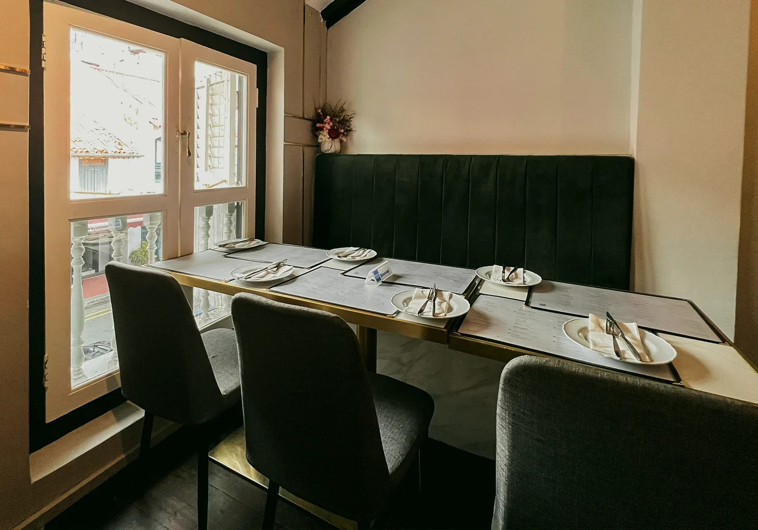 4 The Secret Garden By Zeekri Wide‑angle interior shot of an elegant café dining area with a marble table set for four, plush green upholstered bench seating, minimalist tableware, and natural daylight streaming through tall windows.
