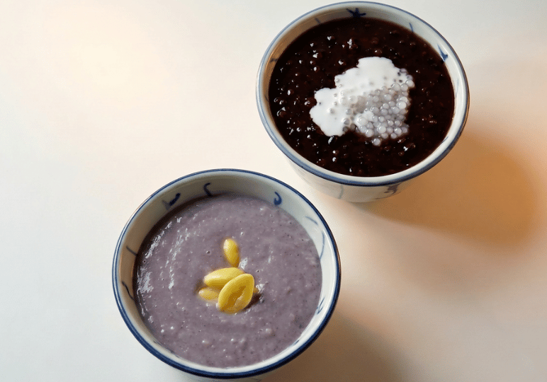 4-curry-times-velocity Two bowls of traditional Singaporean desserts, one containing black glutinous rice with coconut milk and one with a purple bean paste.
