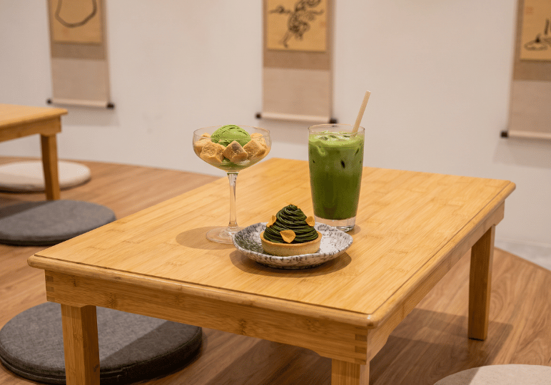 A low wooden table displaying an iced matcha latte, a matcha ice cream and warabimochi dessert in a coupe glass, and an intricate matcha mont blanc tart on a speckled ceramic plate.
