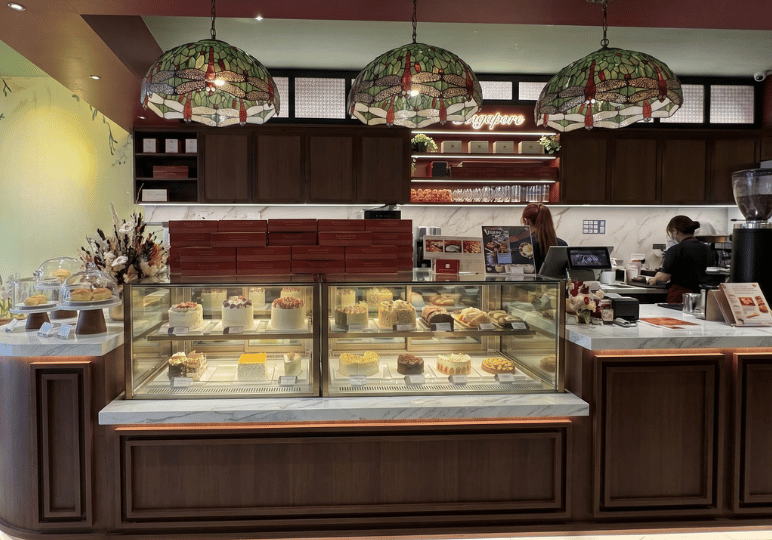 4-restaurant-interior A charming cafe counter displaying various cakes in glass cases, illuminated by three vintage-style stained glass dragonfly pendant lights against a backdrop of warm wood cabinetry.