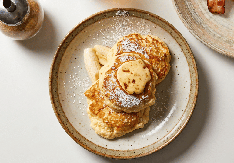 A top-down view of a stack of thick, fluffy pancakes dusted with powdered sugar and topped with a melting pat of butter, served with sliced bananas on a speckled ceramic plate.