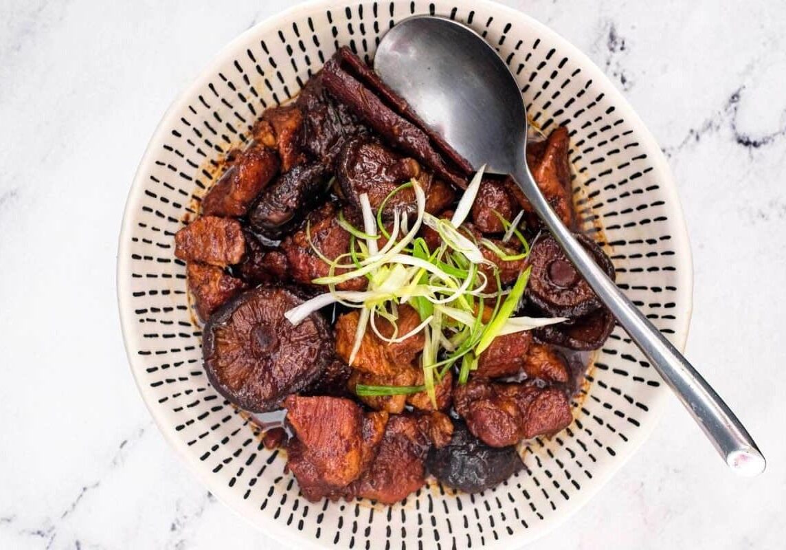 This image displays a bowl of Babi Pongteh, a classic Peranakan braised pork dish featuring tender meat and whole shiitake mushrooms in a savory soybean gravy. The dish is garnished with fresh slivered scallions and presented in a decorative bowl with a dark dash pattern, set against a clean marble backdrop.