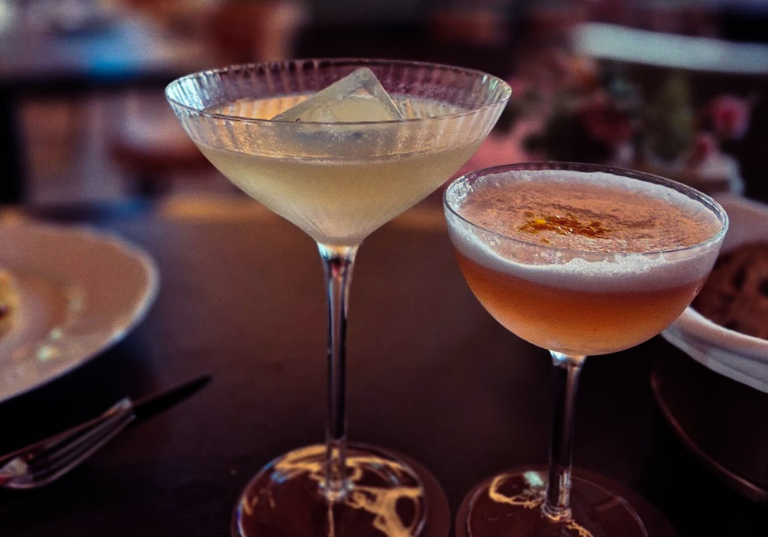 Two elegant cocktails on a dark tabletop — a pale yellow drink with a large clear ice cube and a frothy amber cocktail with garnish, set in a dim restaurant with blurred plates and floral decor.