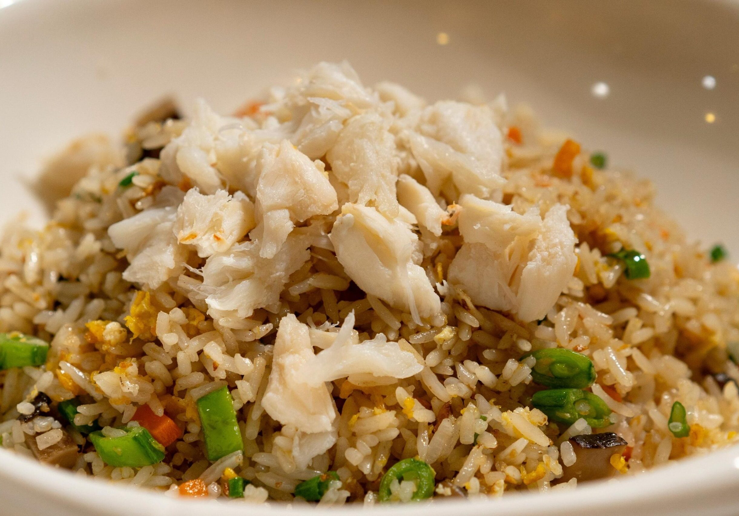 This image showcases a vibrant bowl of crab fried rice, topped with generous chunks of fresh white crab meat. The dish is tossed with colorful ingredients like green onions, carrots, and bits of egg, all served in a clean white ceramic bowl.