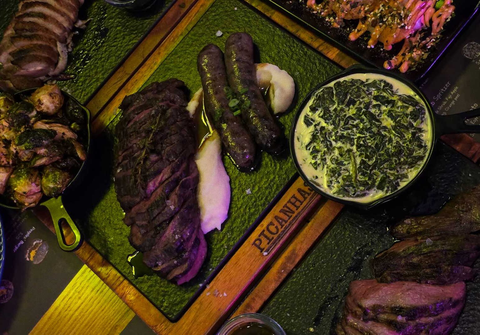 Wooden board labeled Picanhas with sliced steak, sausages on mashed potatoes, bowl of creamed spinach, and side of Brussels sprouts.