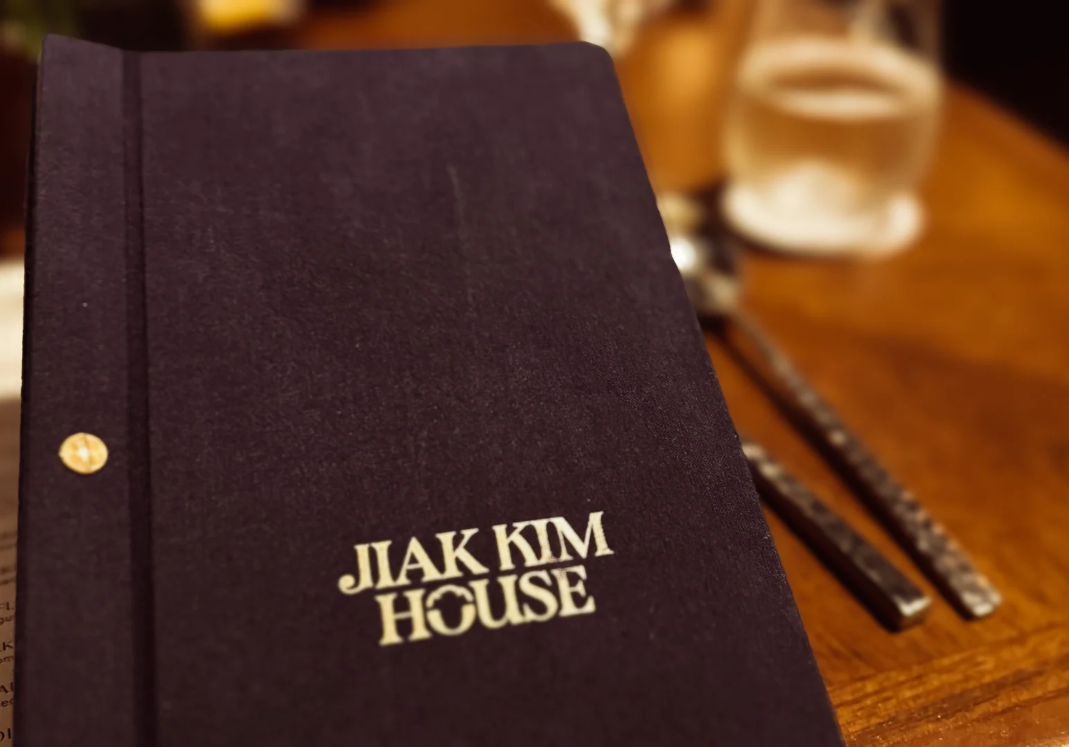 5 Jiak Kim House Singapore Close-up of the Jiak Kim House menu with a textured dark cover resting on a wooden dining table beside cutlery and glassware.