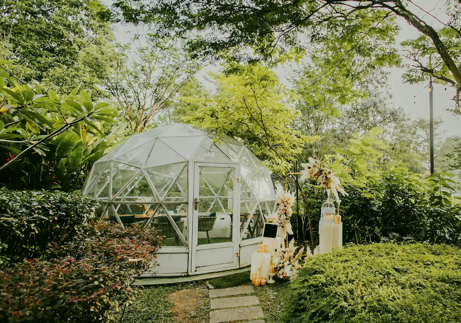 Eye‑level wide shot of a glass dome dining setup surrounded by lush greenery, featuring a private romantic table arrangement in a garden setting.