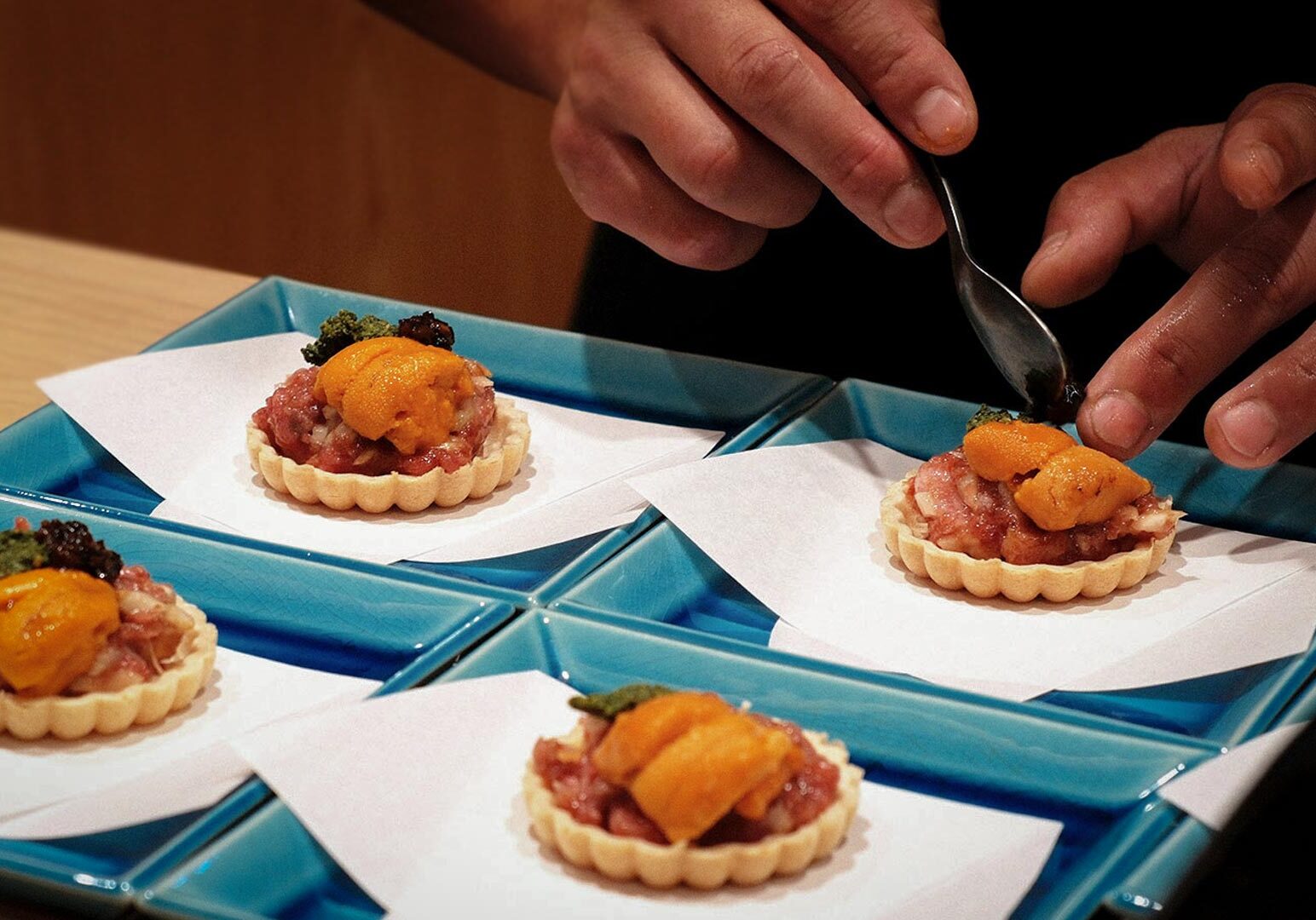 Close‑up of gourmet tartlets topped with tuna, sea urchin (uni), caviar, and garnish, plated on blue dishes in fine dining setting.