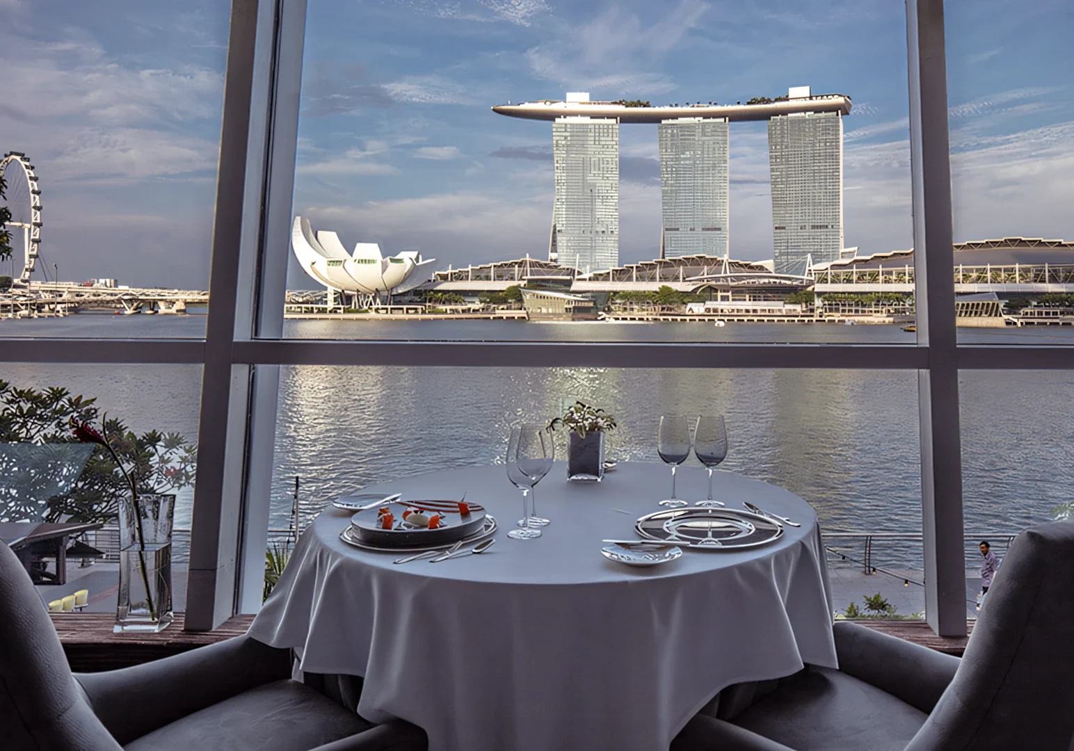 Table set for two overlooking the Marina Bay Sands skyline, with wine glasses, plates, and a waterfront cityscape through floor‑to‑ceiling windows.