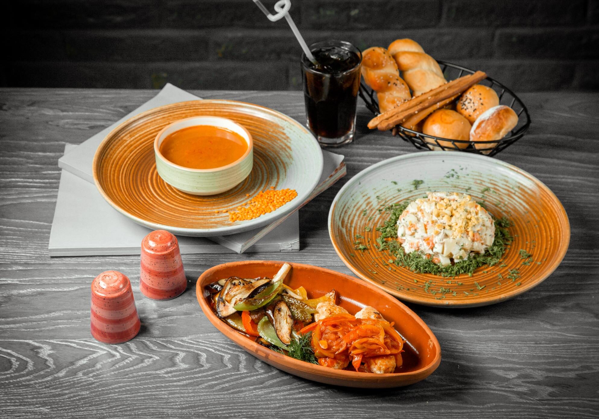 This image presents an elaborate meal set on a grey wooden table, featuring a bowl of orange soup, a creamy salad garnished with dill, and an oval dish of meatballs with sautéed vegetables. The spread is rounded out by a basket of assorted breads, a cold dark soda with a straw, and matching red salt and pepper shakers.