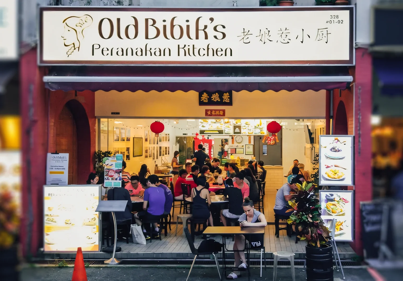 Old Bibik’s Peranakan Kitchen storefront with illuminated signage, outdoor seating, food display boards, and diners enjoying meals inside the brightly lit restaurant.