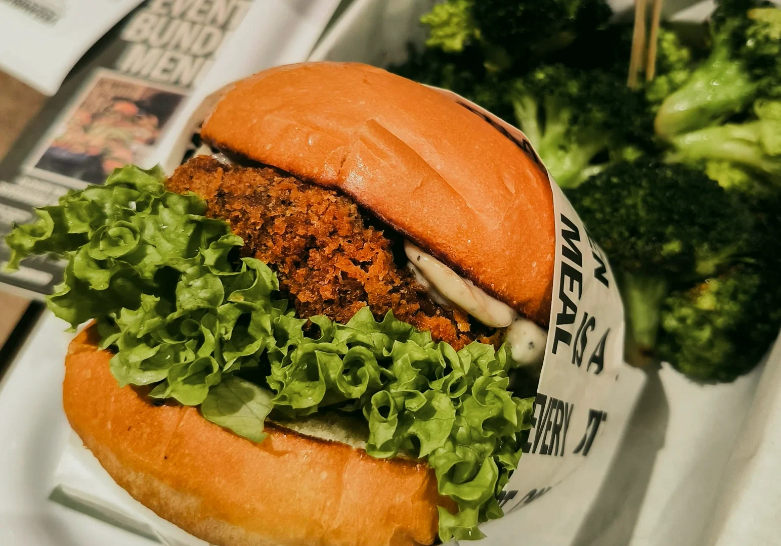 Close‑up three‑quarter angle shot of a vegetarian burger with a crispy plant‑based patty, fresh green lettuce, soft toasted bun, and broccoli side dish served on a white plate.