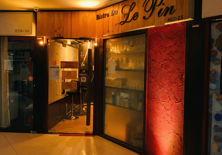 The exterior entrance of Bistro du Le Pin located at unit #02-25 inside Orchard Plaza, showcasing a wooden sign and a distinctive red textured wall.