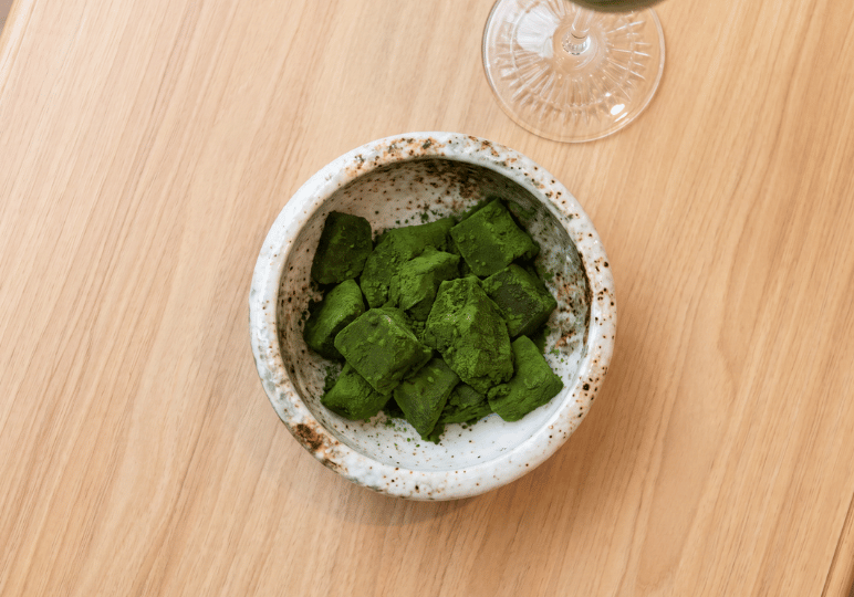 A top-down view of a rustic, white speckled ceramic bowl filled with bright green, matcha-powder-coated warabimochi cubes resting on a light wood table.