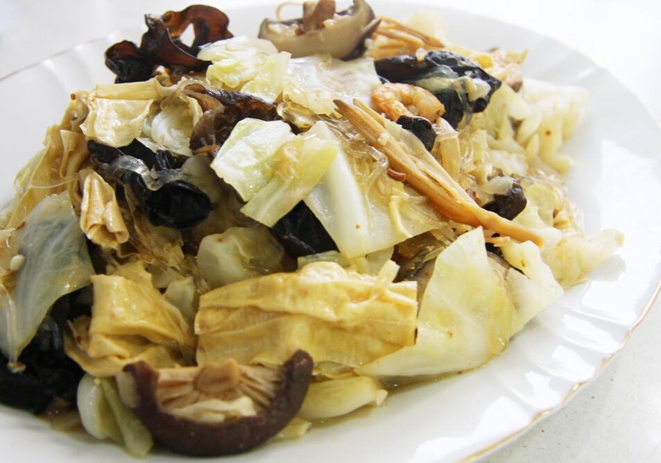 This image features a plate of Nyonya Chap Chye, a traditional Peranakan vegetable stew consisting of cabbage, glass noodles, and lily buds. The dish is further enriched with dark wood ear mushrooms, shiitake mushrooms, and beancurd skin, all served on a simple white scalloped plate.