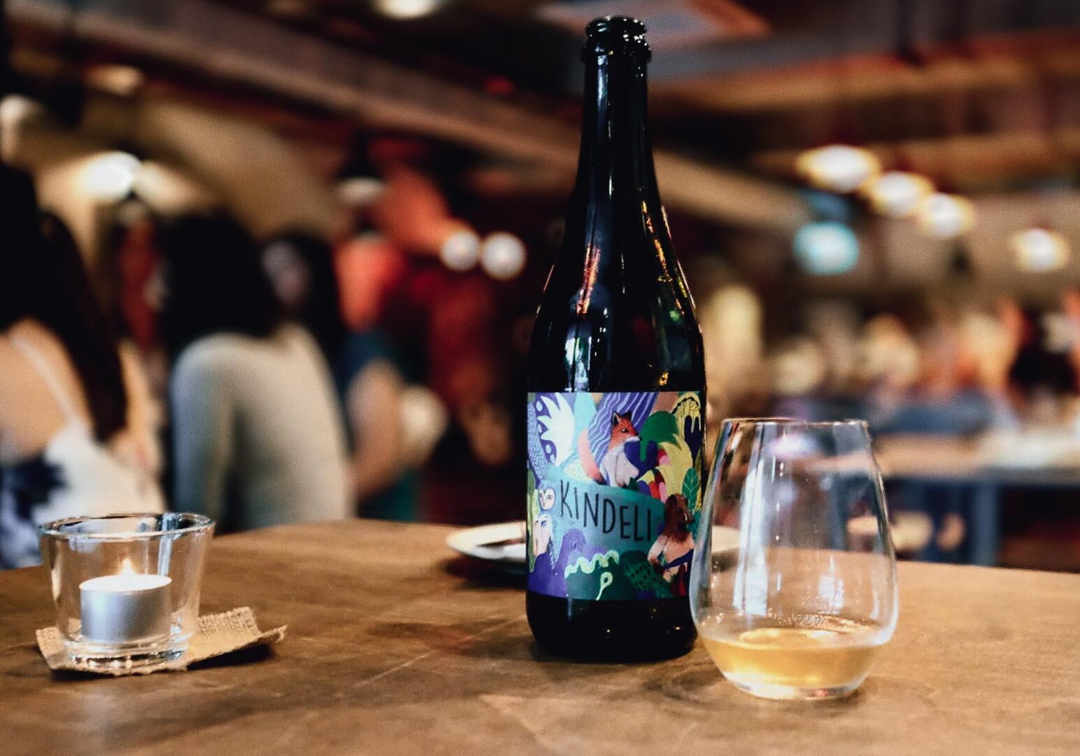 Wine bottle displayed on a wooden table alongside a half‑filled wine glass and a lit tealight candle, set against the warm, blurred ambiance of a lively restaurant or bar.