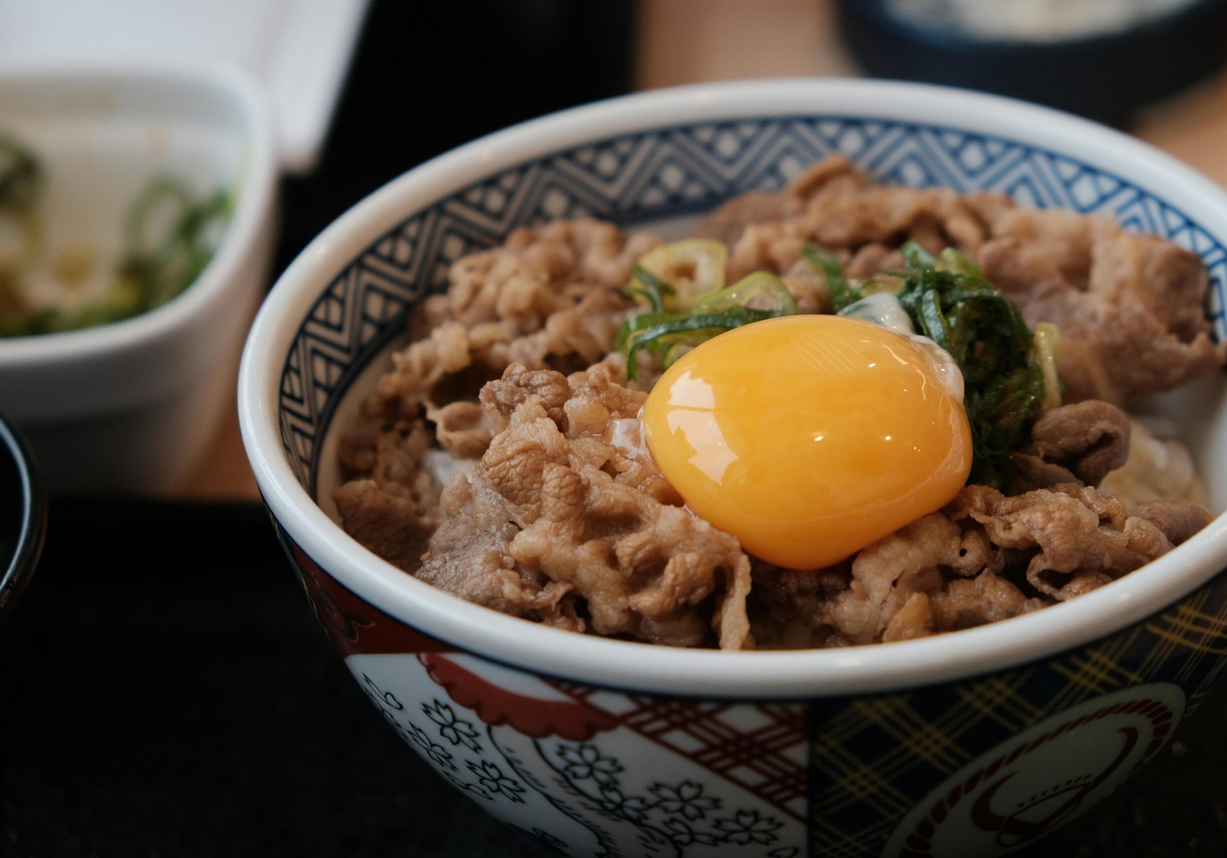 A ceramic bowl with a blue geometric pattern is filled with white rice topped with thinly sliced beef, green onions, and a single raw egg yolk. The bowl sits on a dark table, with blurred side dishes visible in the background of the soft-focus shot.