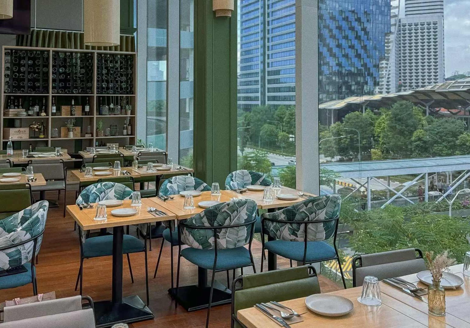 A modern restaurant interior with wooden tables, patterned cushioned chairs, and neatly arranged table settings. Floor‑to‑ceiling windows provide a view of city buildings, greenery, and elevated walkways outside, resembling the Suntec City area.