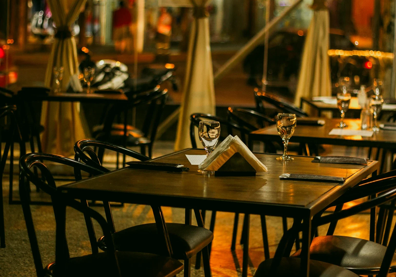Eye-level wide shot of an empty outdoor restaurant dining area at night, with neatly set tables, wine glasses, folded napkins, and warm ambient lighting, reflecting a refined Michelin-style dining atmosphere.