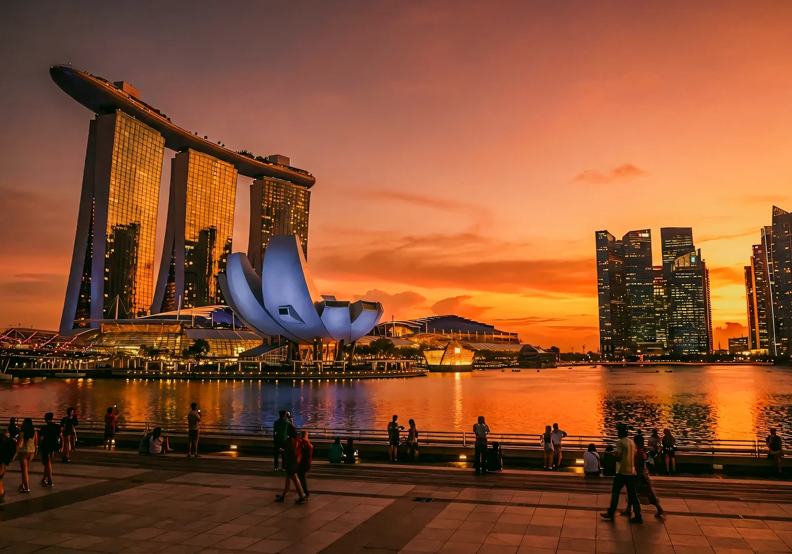 Wide‑angle waterfront cityscape of Marina Bay Sands and the ArtScience Museum in Singapore at sunset, photographed from a low eye‑level perspective along the bay promenade, with illuminated skyscrapers reflecting on calm water and people silhouetted in the foreground.