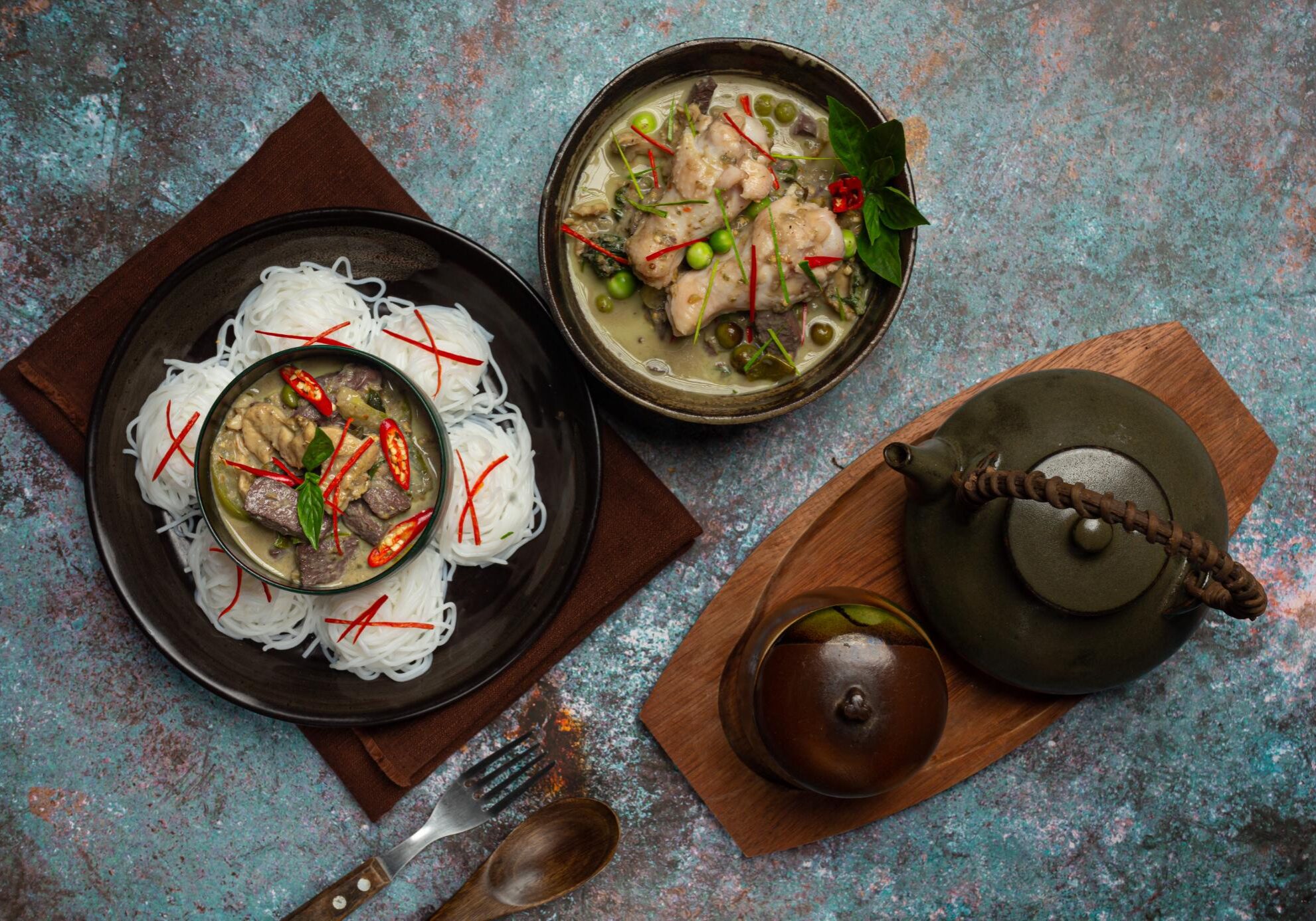 This image showcases a top-down view of a Thai meal featuring two bowls of green curry with chicken and pea aubergines, one of which is served atop nests of thin rice noodles. The spread is arranged on a textured, dark tabletop and includes a rustic clay teapot on a wooden board alongside a dark napkin and wooden cutlery.