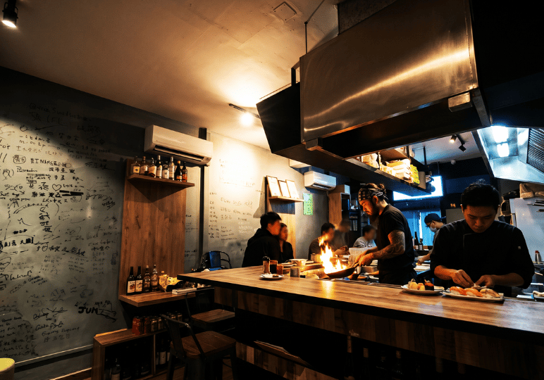 6-orchard-plaza-stalls A tattooed chef cooking with a large flame behind the wooden counter of an intimate izakaya, featuring a wall covered in handwritten notes and signatures from guests.