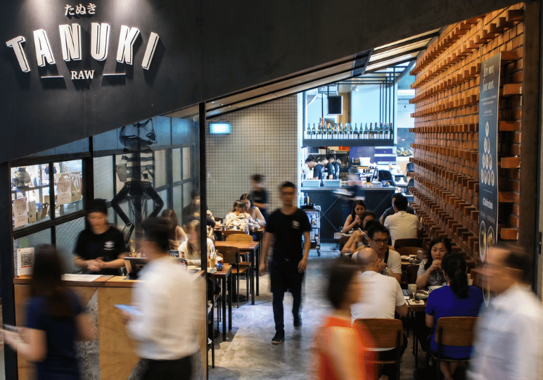 The busy storefront and dining area of Tanuki Raw, featuring a prominent black sign, brick wall accents, and several tables filled with patrons.
