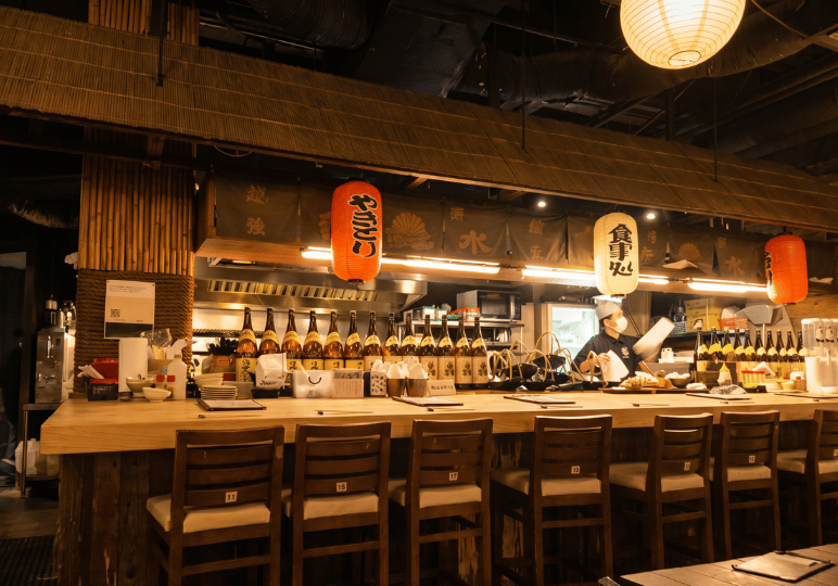 Traditional Japanese izakaya-style bar counter seating at Tomo Tokyo, featuring wooden stools, rows of sake bottles, and hanging paper lanterns over the kitchen pass.